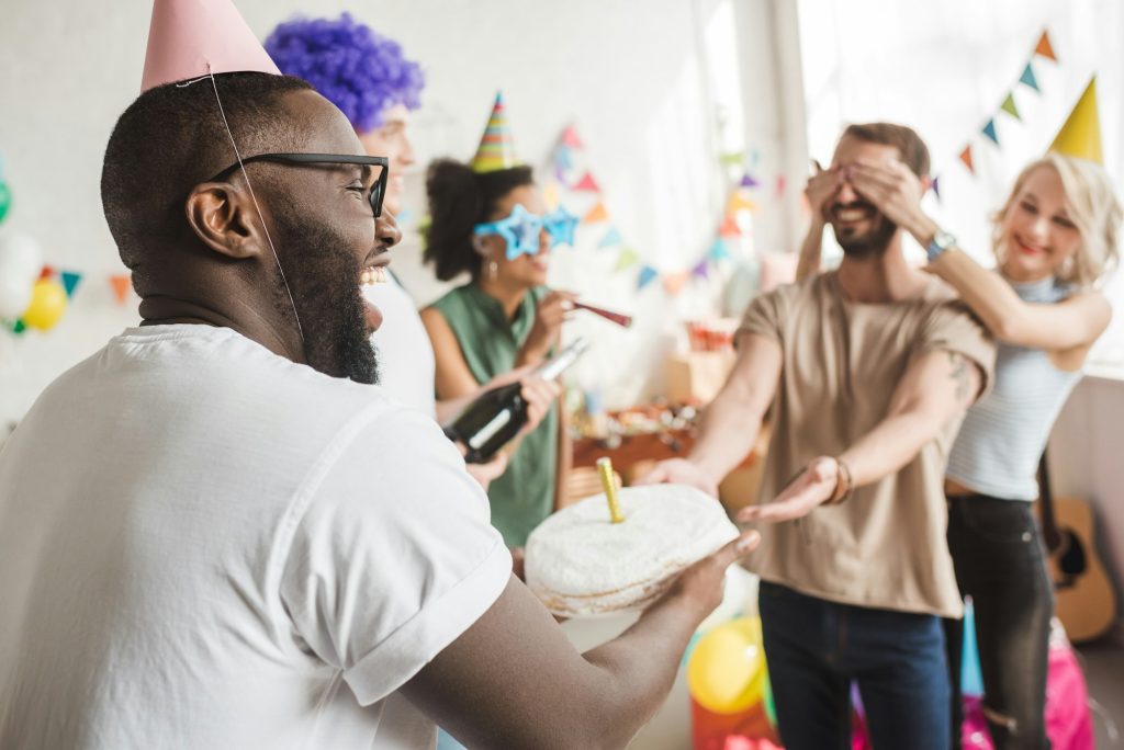 Happy friends covering eyes of young man and greeting him with birthday cake